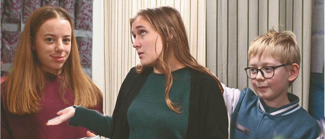 Drei Personen stehen vor einem gestreiften Vorhang. Eine Frau hält eine Geschenkbox und ein Mädchen lächelt sie an. Ein Junge mit Brille schaut interessiert zu.