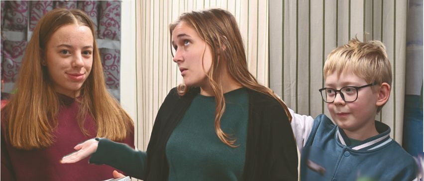 Drei Personen stehen vor einem gestreiften Vorhang. Eine Frau hält eine Geschenkbox und ein Mädchen lächelt sie an. Ein Junge mit Brille schaut interessiert zu.