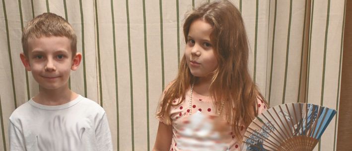 Zwei Kinder, ein Junge und ein Mädchen, stehen eng beieinander. Der Junge trägt ein weißes T-Shirt und das Mädchen hält einen Fächer. Hinter ihnen befindet sich ein Vorhang mit grünen Streifen.