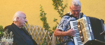 Two men sit on a stone patio; one plays an accordion while the other listens, surrounded by greenery and a stone wall.