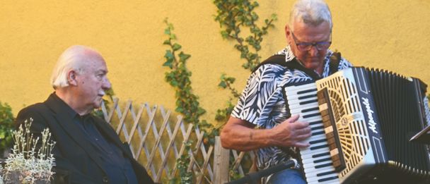 Zwei Männer sitzen auf einer Steinterrasse; einer spielt Akkordeon, während der andere zuhört, umgeben von Grünpflanzen und einer Steinmauer.