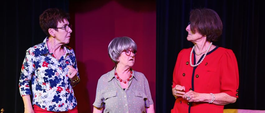 Drei ältere Frauen stehen auf einer Bühne in einem Theater und tragen verschiedene Outfits und Accessoires. Die Frau auf der linken Seite trägt eine Brille und rote Hosen. Die Frau in der Mitte trägt ein graues Kleid mit einer Halskette. Die Frau auf der rechten Seite trägt ein rotes Kleid mit einer Perlenkette.