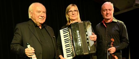 Drei Personen stehen auf einer Bühne, zwei Männer und eine Frau. Der Mann links hält eine Flasche, die Frau in der Mitte ein Akkordeon und der Mann rechts eine Geige. Sie sind alle formell gekleidet.