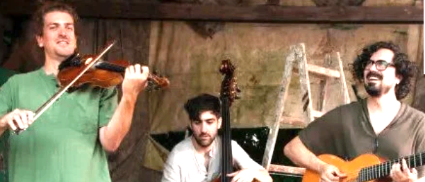 Drei Männer in lässiger Kleidung spielen Musikinstrumente. Der Mann auf der linken Seite spielt Geige, der Mann in der Mitte spielt Cello und der Mann auf der rechten Seite spielt Gitarre. Sie stehen in einer Outdoor-Umgebung.