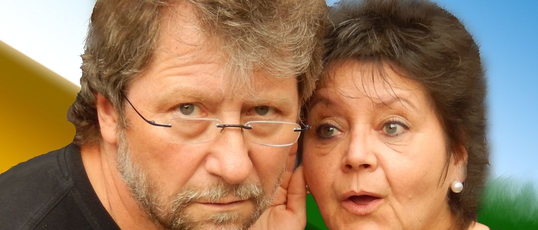 Ein Mann und eine Frau stehen eng beieinander, beide tragen schwarze T-Shirts. Der Mann hat eine Brille und einen Bart, die Frau hat kurze Haare. Sie posieren für ein Foto.