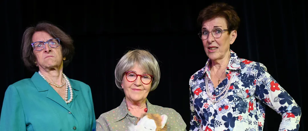 Drei ältere Frauen stehen zusammen, eine hält eine Plüschkatze. Sie sind in verschiedenen farbigen Kleidungsstücken gekleidet und scheinen auf der Bühne zu stehen.