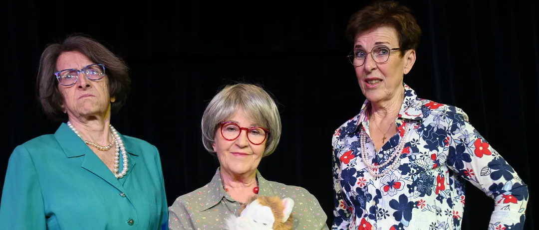 Drei ältere Frauen stehen zusammen, eine hält eine Stoffkatze, alle tragen Brillen und lächeln für die Kamera.