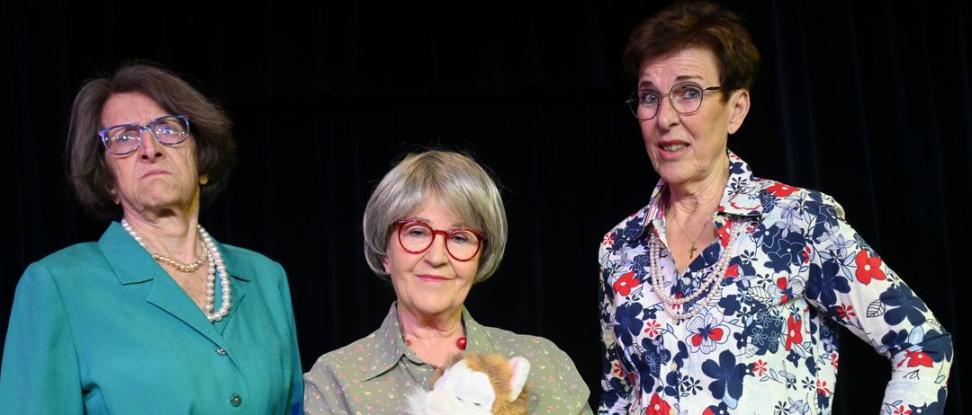Drei ältere Frauen stehen zusammen, eine hält eine Stoffkatze, alle tragen Brillen und lächeln für die Kamera.