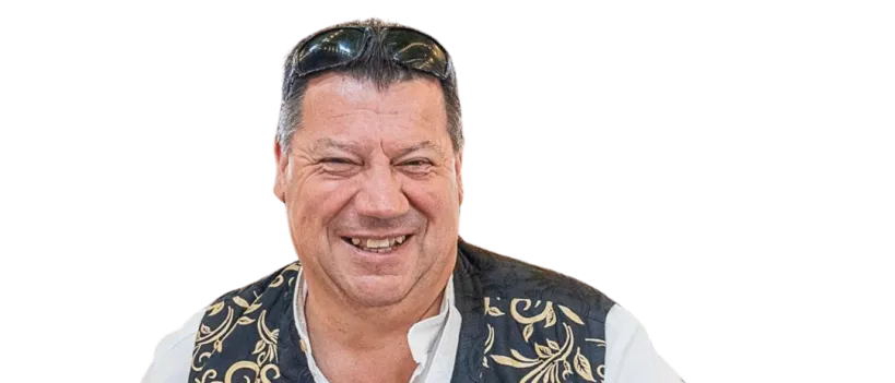 Ein Mann mit einem lächelnden Gesicht, der ein weißes Hemd und eine schwarz-goldene Weste mit verschiedenen Abzeichen trägt und eine Sonnenbrille auf dem Kopf hat.