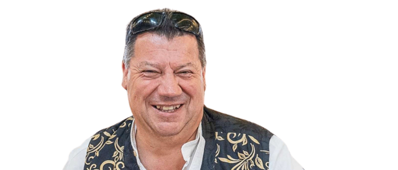 Ein Mann mit einem lächelnden Gesicht, der ein weißes Hemd und eine schwarz-goldene Weste mit verschiedenen Abzeichen trägt und eine Sonnenbrille auf dem Kopf hat.