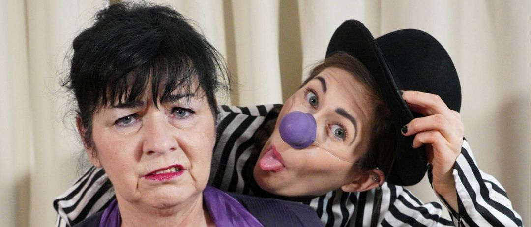 Drei Frauen in einer theatralischen Pose. Die Frau in der Mitte lacht, während die Frau auf der linken Seite streng aussieht. Die Frau auf der rechten Seite hat eine lila Nase und trägt ein gestreiftes Hemd. Der Text lautet 'Nichts ist wie es scheint.'