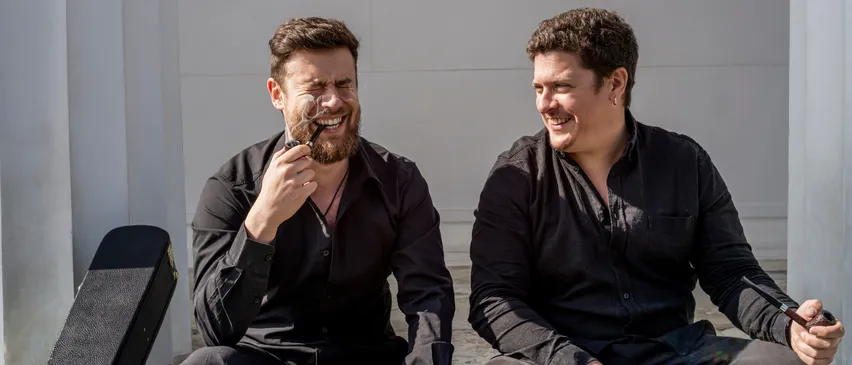 Zwei Männer in schwarzen Hemden sitzen auf einer Betonbank, einer lächelt und hält eine Pfeife, der andere lacht mit einem Gitarrenkoffer in der Nähe.
