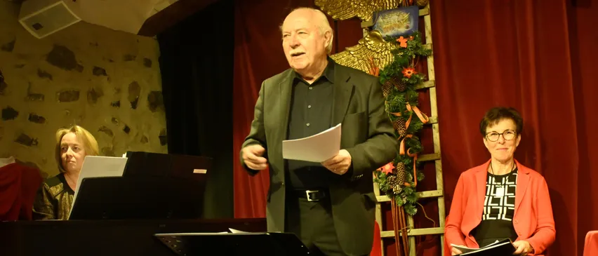 Ein älterer Mann steht auf der Bühne und hält ein Stück Papier in der Hand, wahrscheinlich hält er eine Rede. Hinter ihm ist ein Klavier zu sehen, und eine Frau sitzt auf einem Stuhl. Eine Leiter mit Weihnachtsdekorationen ist im Hintergrund.