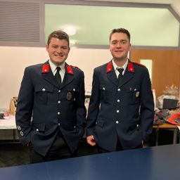 Zwei Männer in Uniform stehen nebeneinander und lächeln für ein Foto. Sie stehen vor einem Tisch und einer Tür, auf dem Tisch befinden sich verschiedene Gegenstände.