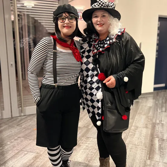 Zwei Frauen in Clownkostümen stehen eng nebeneinander in einem Gebäudeflur. Eine trägt ein gestreiftes Shirt, Hosenträger und Socken, die andere eine karierte Jacke und einen Hut.