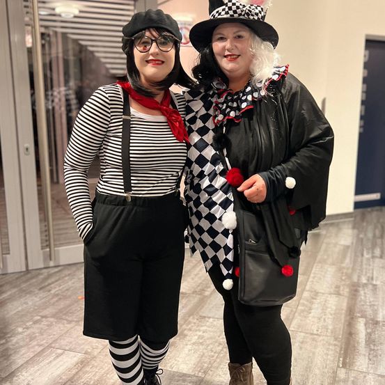 Zwei Frauen in Clownkostümen stehen eng nebeneinander in einem Gebäudeflur. Eine trägt ein gestreiftes Shirt, Hosenträger und Socken, die andere eine karierte Jacke und einen Hut.