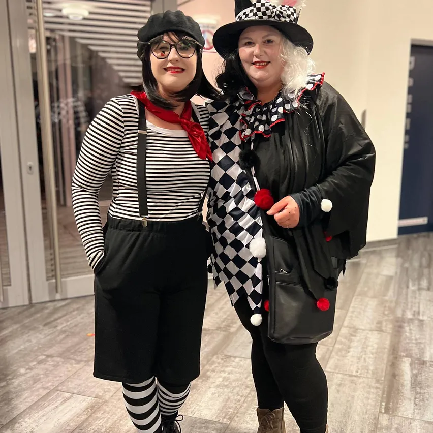 Zwei Frauen in Clownkostümen stehen eng nebeneinander in einem Gebäudeflur. Eine trägt ein gestreiftes Shirt, Hosenträger und Socken, die andere eine karierte Jacke und einen Hut.