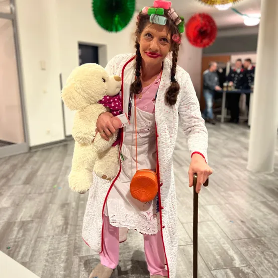 Eine Frau, die als Krankenschwester gekleidet ist, hält einen weißen Teddybären und eine Handtasche. Sie steht in einem Flur.
