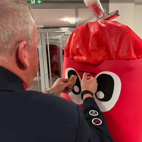 Eine uniformierte Person steckt Geld in einen roten, charakterbasierten Spendenbehälter mit einem Plastikbeutel obenauf.