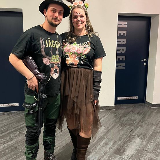 Ein Mann und eine Frau in Hirschkostümen, stehen zusammen und lächeln für die Kamera. Der Mann hält eine Spielzeugpistole, und die Frau hat Blumen in den Haaren. Hinter ihnen befindet sich eine Tür mit der Aufschrift 'Herren'.