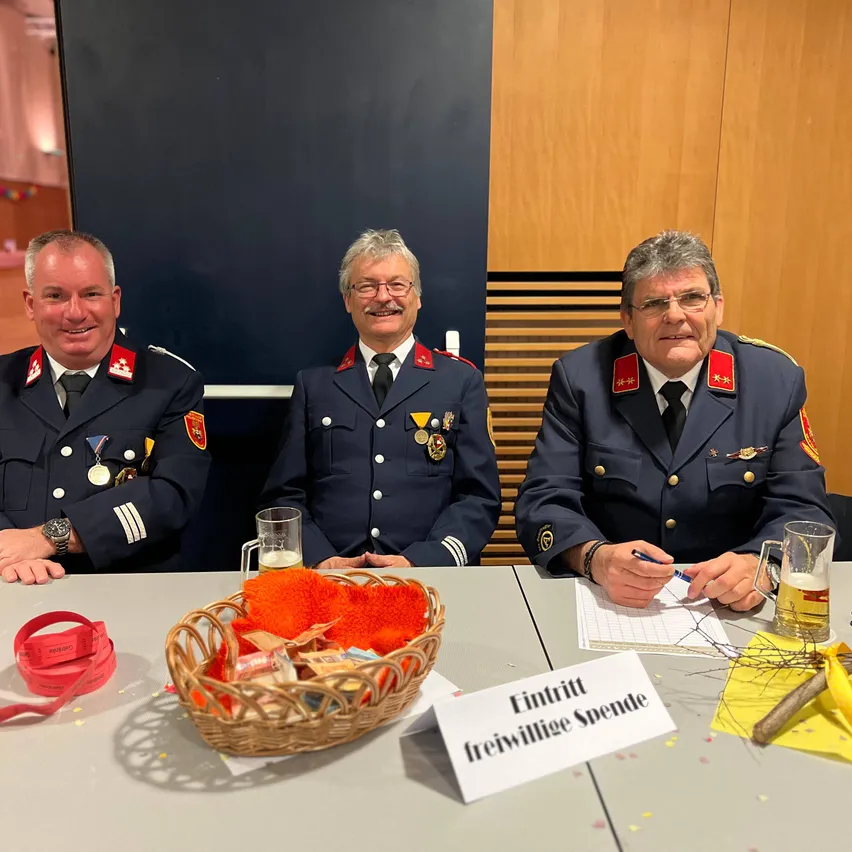 Drei Männer in Uniformen sitzen an einem Tisch mit einem Korb, Bierkrügen und Papieren. Einer schreibt, während andere lächeln. Ein Schild steht 'Eintritt freiwillige Spende'.
