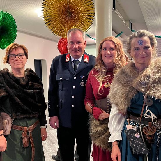 Vier Personen stehen zusammen drinnen. Ein Mann in Uniform, eine Frau mit roten Haaren und zwei weitere in Pelz- und historischer Kleidung. Hinter ihnen hängen dekorative Papierfächer.