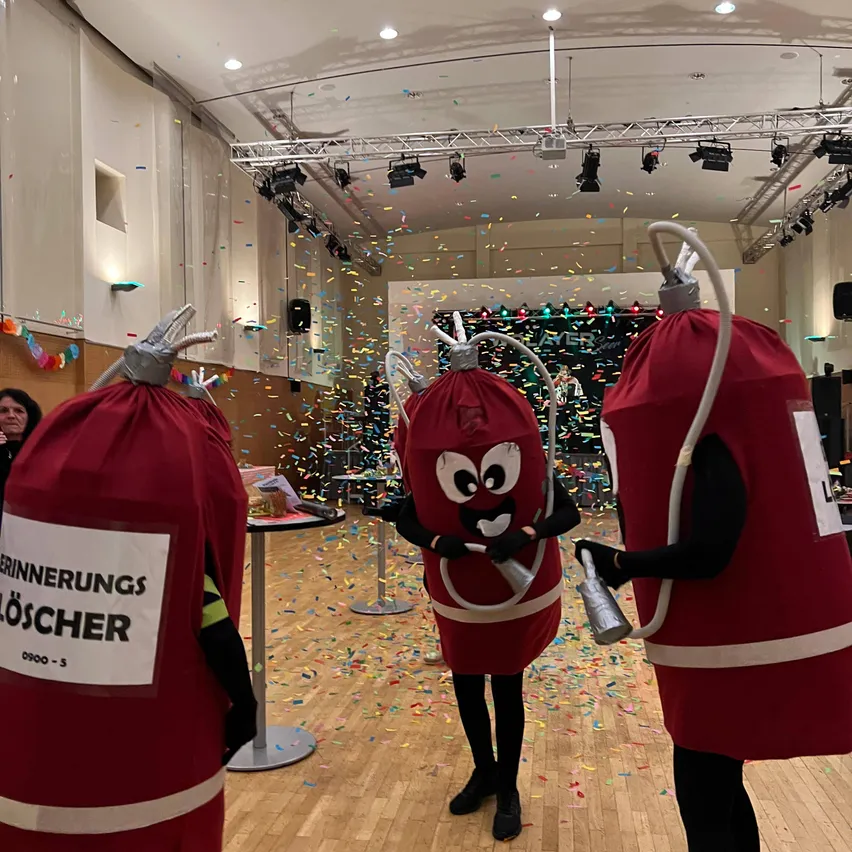 Drei Personen in roten Feuerwehrmann-Kostümen stehen in einem mit Konfetti gefüllten Raum. Einer hält eine Glocke.