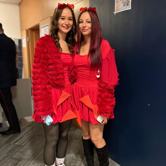 Zwei Frauen in roten Kostümen, eine mit einem Pelzmantel und die andere mit einem federnden Rock, posieren für ein Foto. Beide haben rote Bögen auf dem Kopf. Sie stehen in einem Flur mit einer blauen Wand.