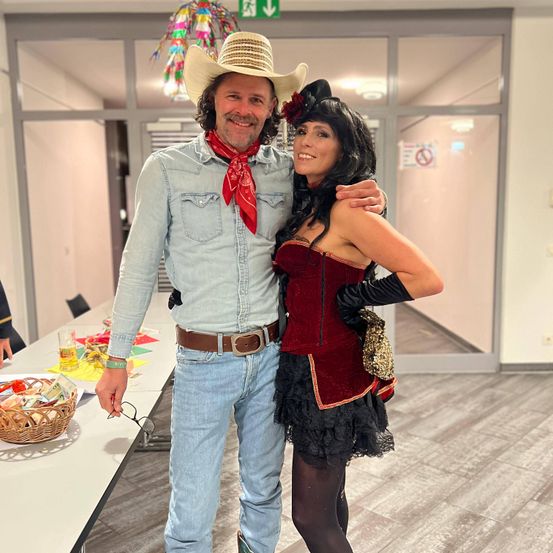 Ein Mann und eine Frau in Kostümen stehen lächelnd eng beieinander. Sie stehen in einem Saal mit Glastüren.