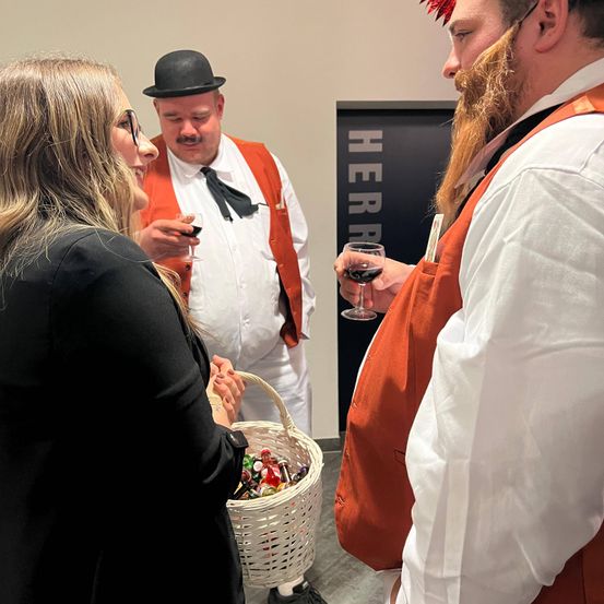 Eine Frau mit Brille hält einen Korb mit Süßigkeiten und unterhält sich mit zwei Männern in orangefarbenen Westen, einer hält ein Weinglas. Dahinter steht ein Schild mit der Aufschrift 'Herr'.