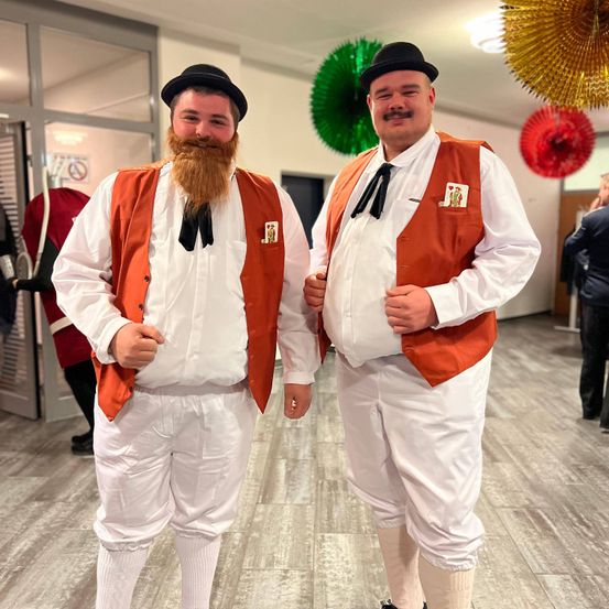 Zwei Männer in übereinstimmenden orangefarbenen Westen und weißen Hemden posieren in einer Halle. Sie tragen Hüte und Fliegen. Bunte Papierfächer hängen oben.