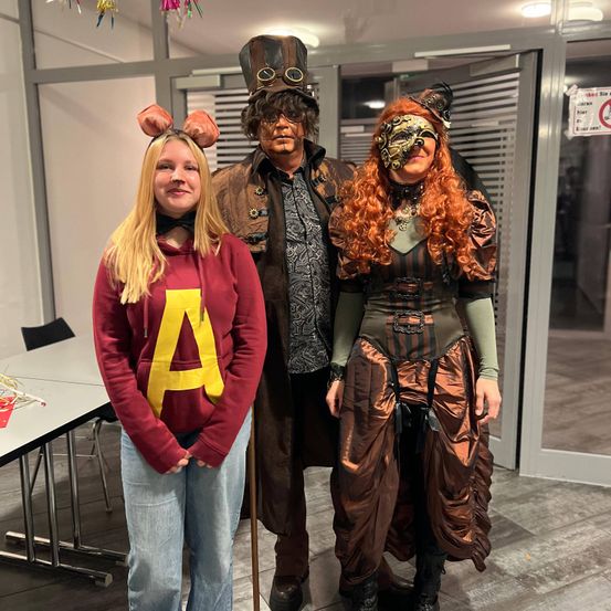 Drei Menschen in Kostümen posieren für ein Foto. Eine Frau in einem roten Sweatshirt mit dem Buchstaben A, ein Mann in einem Steampunk-Hut und -Mantel und eine Frau in einem Steampunk-Kleid und -Maske.