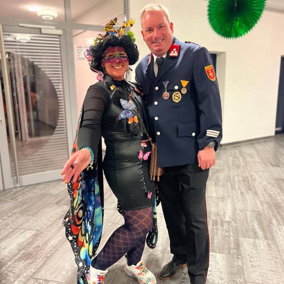 Eine Frau in einem Schmetterlings-Kostüm steht neben einem dienstlich gekleideten Mann mit Medaillen auf der Brust. Sie posieren für ein Foto in einem gut beleuchteten Raum.