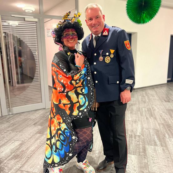 Ein Mann in Uniform steht neben einer Frau in einem Schmetterlingskostüm. Sie lächeln und posieren für ein Foto. Hinter ihnen ist eine grüne kreisförmige Dekoration.