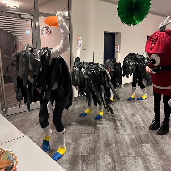 Mehrere Menschen in Kostümen, die Straußen ähneln, stehen in einem Flur. Sie halten orangefarbene Objekte. Der Flur hat eine Glastür, eine Tür und eine grüne kreisförmige Dekoration.