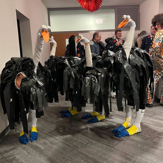 Zwei Personen in Straußenkostümen mit Plastikfolie und weißen Handschuhen stehen in einem Innenbereich. Hinter ihnen stehen einige Leute und unterhalten sich. Eine Person in der rechten Ecke hält ein Glas mit Getränk.