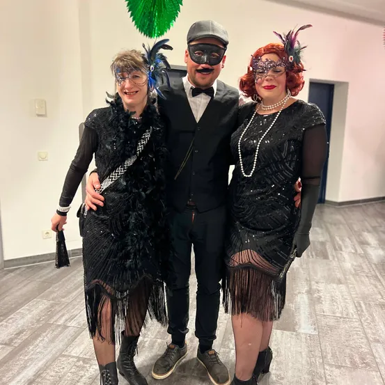 Drei Personen in einem Raum, zwei Frauen und ein Mann, alle in 1920er-inspirierten Kostümen, posieren für ein Foto. Der Mann trägt eine Maske und eine Fliege.