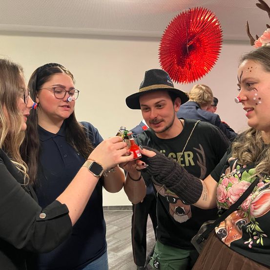 Drei Personen in einem Raum, zwei Frauen und ein Mann. Die Frauen tragen Gesichtsbemalung und halten rote Flaschen. Der Mann trägt einen Hut und ein hirschartiges T-Shirt. Sie scheinen sich auf eine Feier vorzubereiten.