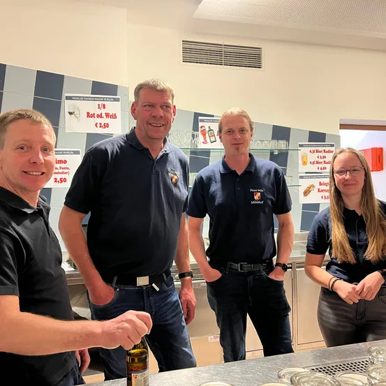 Vier Menschen stehen in einer Küche und lächeln. Sie tragen alle blaue Poloshirts. Hinter ihnen ist eine Wand mit Schildern und einem Regal mit Gläsern.