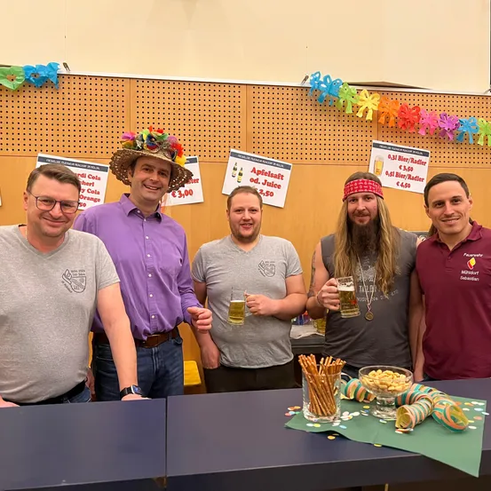 Fünf Männer stehen hinter einem Tisch und halten Biergläser in der Hand. Der Mann in der Mitte trägt einen blumigen Hut, und ein Schild an der Wand hinter ihnen bewirbt Apfelsaft und andere Getränke.