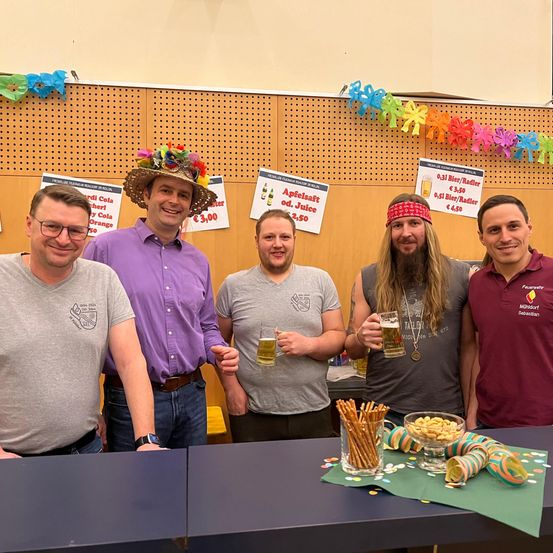 Fünf Männer stehen hinter einem Tisch und halten Biergläser in der Hand. Der Mann in der Mitte trägt einen blumigen Hut, und ein Schild an der Wand hinter ihnen bewirbt Apfelsaft und andere Getränke.