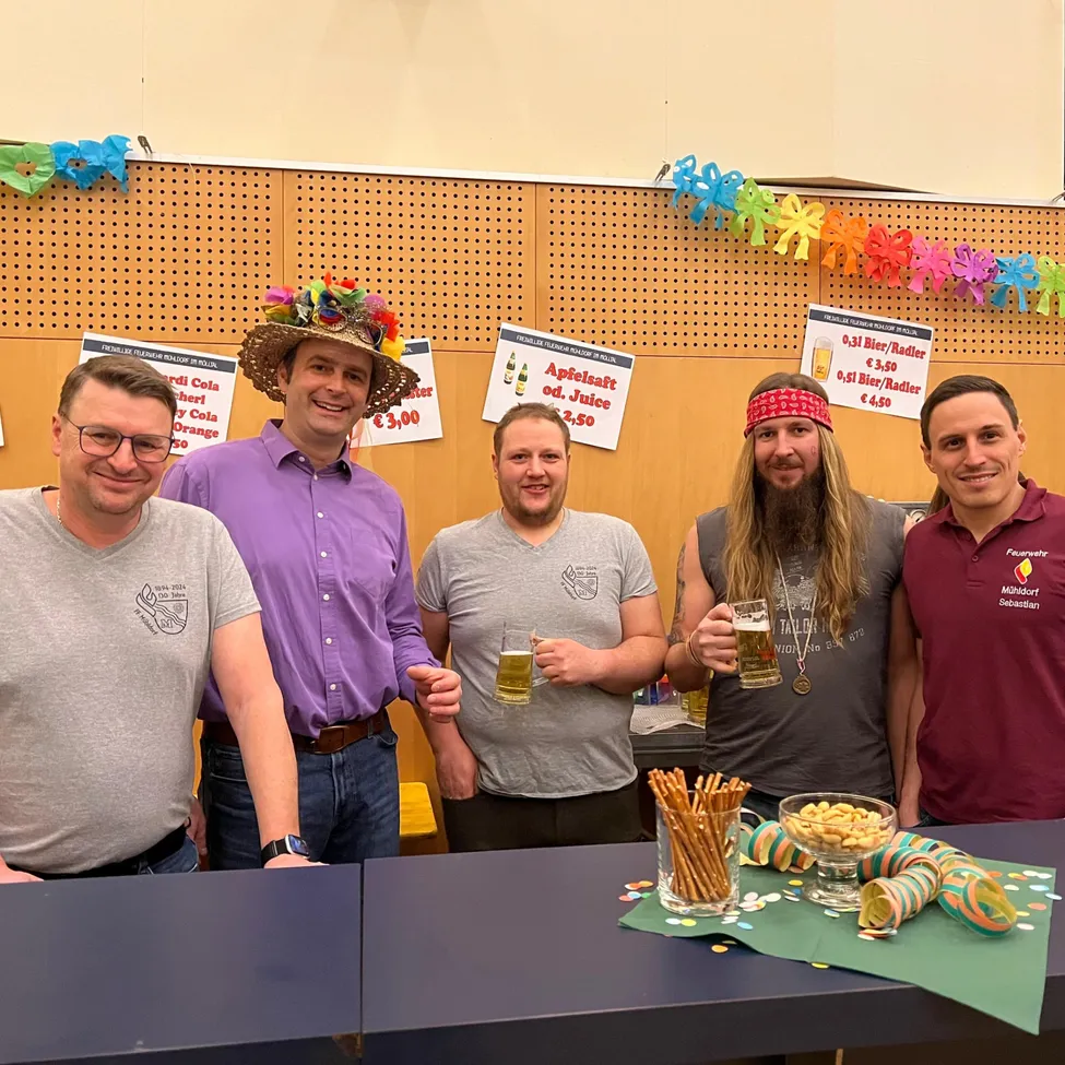 Fünf Männer stehen hinter einem Tisch und halten Biergläser in der Hand. Der Mann in der Mitte trägt einen blumigen Hut, und ein Schild an der Wand hinter ihnen bewirbt Apfelsaft und andere Getränke.