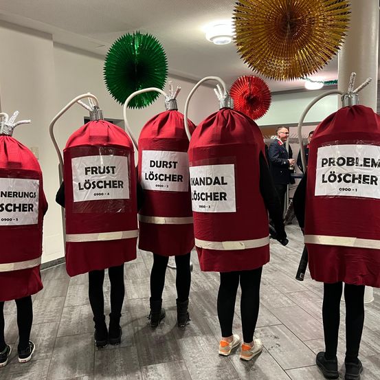 Fünf Personen in roten Kostümen, die Feuerlöschern ähneln, stehen in einem Raum. Jedes Kostüm hat ein Etikett mit Namen und Nummer. Ein Mann in einem Anzug steht in der Nähe.