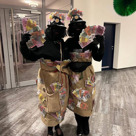 Zwei Personen in Schwarz mit Juteschurze, geschmückt mit vielen Euro-Scheinen, posieren drinnen in der Nähe einer Glastür.