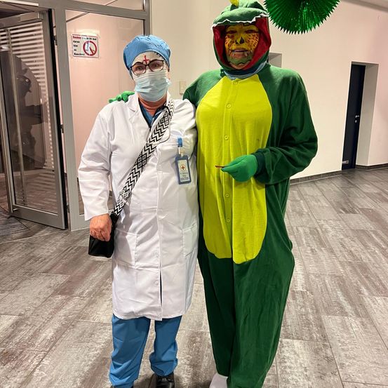 Eine Person in einem Arztkittel und eine Person in einem Krokodilkostüm stehen in einem Gebäude.