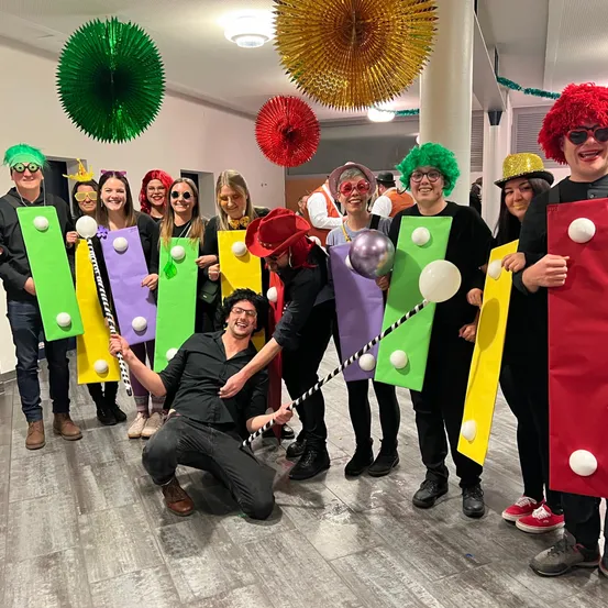 Eine Gruppe von Menschen in Kostümen posiert für ein Foto in einem dekorierten Raum mit Ballons und Schildern.