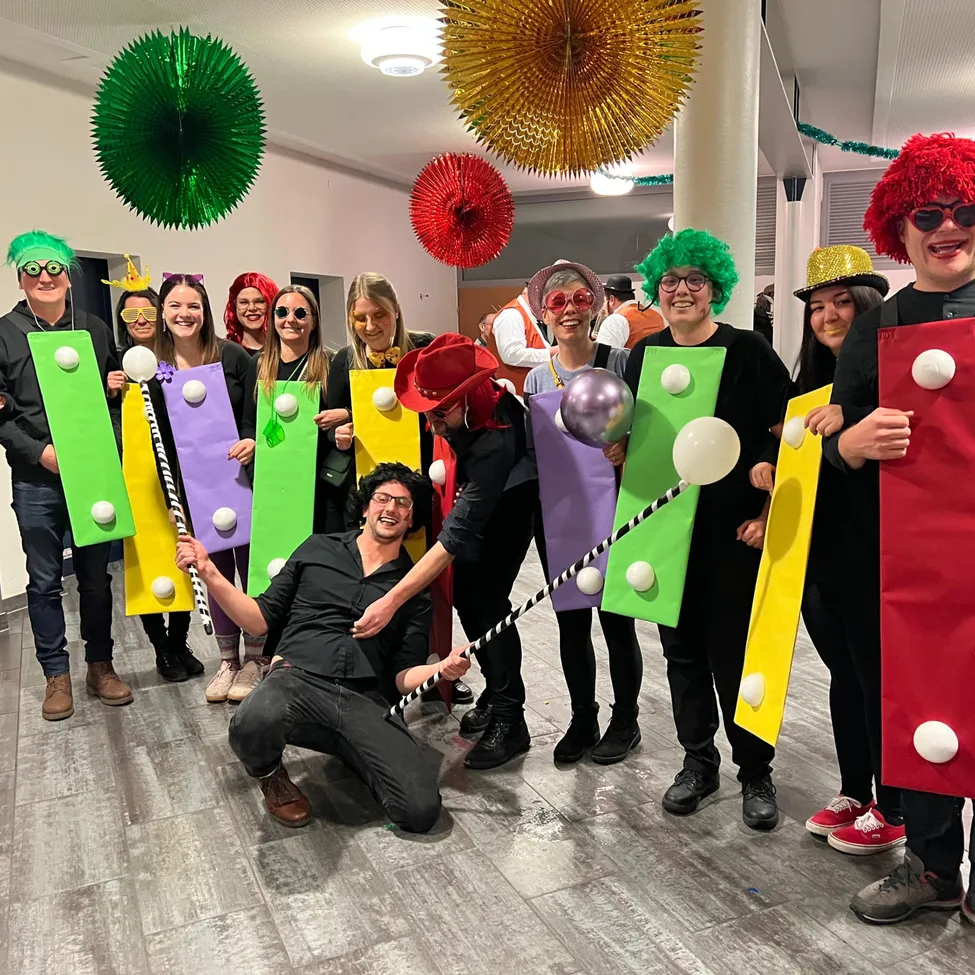 Eine Gruppe von Menschen in Kostümen posiert für ein Foto in einem dekorierten Raum mit Ballons und Schildern.