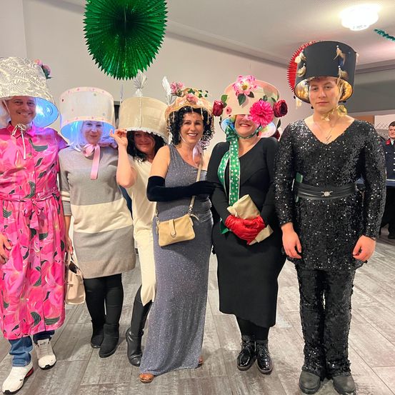 Eine Gruppe von Menschen in schicken Hüten posiert für ein Foto. Sie tragen verschiedene Outfits, und einige tragen Handschuhe. Eine von ihnen hält eine Handtasche. Dahinter ist ein verschwommener Blick auf einen grünen Fächer und eine Person in Uniform.