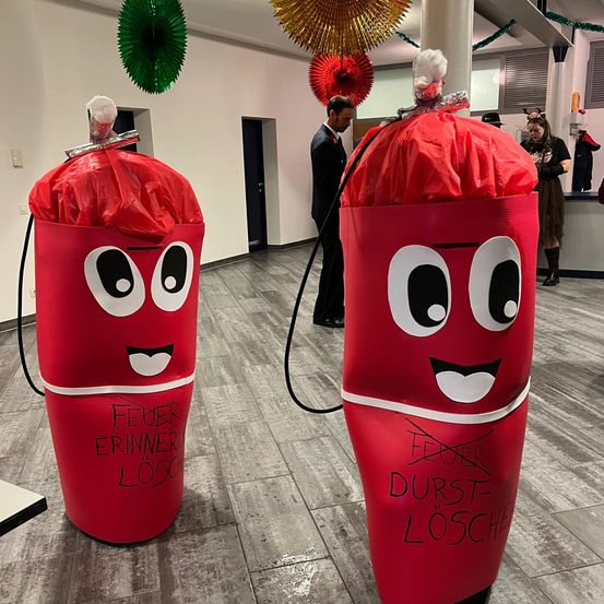 Zwei rote Feuerlöscher stehen auf einem gefliesten Boden. Jeder hat ein Gesicht mit Augen und Mund. Einer steht 'Feuer Erinnerung Löschen' und der andere 'Feuer Durst Löschen.' Ein Mann in Anzug steht dahinter.