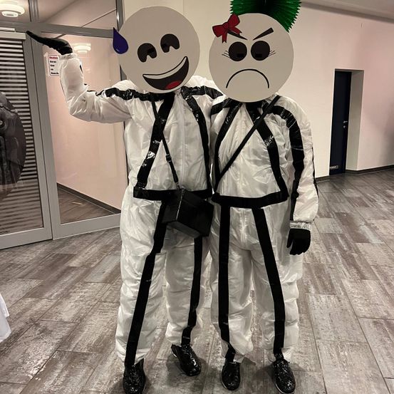 Zwei Personen, die weiße Anzüge mit schwarzen Streifen und Gesichtsmasken mit einem lächelnden und einem wütenden Gesicht tragen, stehen in einem Flur.